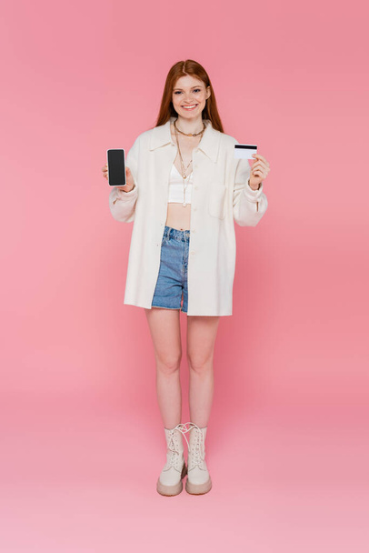 Kaunis ja tyylikäs punatukkainen nainen, jolla on matkapuhelin ja luottokortti vaaleanpunaisella taustalla - Valokuva, kuva