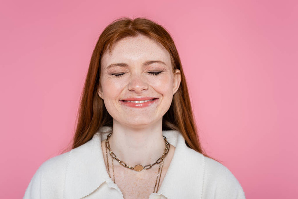 Mujer pecosa feliz cerrando los ojos y sonriendo aislado en rosa  - Foto, imagen