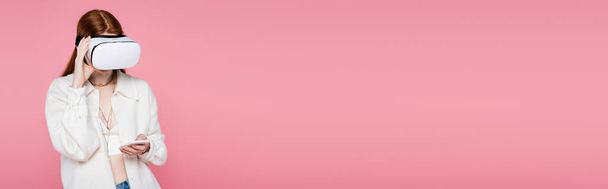 Jovem mulher ruiva no fone de ouvido vr usando smartphone isolado em rosa, banner  - Foto, Imagem