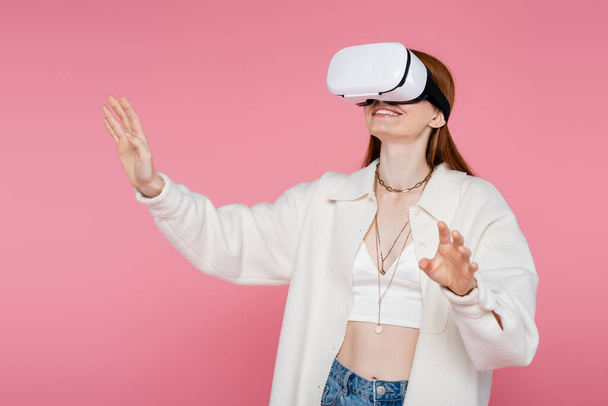 Cheerful and trendy woman in jacket gaming in vr headset isolated on pink  - 写真・画像