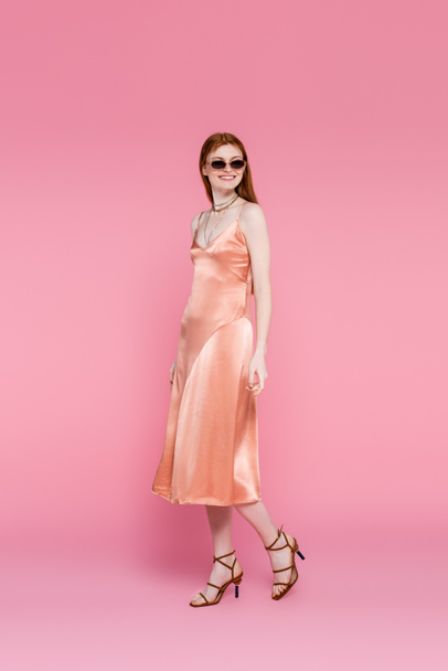 Smiling woman in silk dress and sunglasses walking on pink background  - Valokuva, kuva