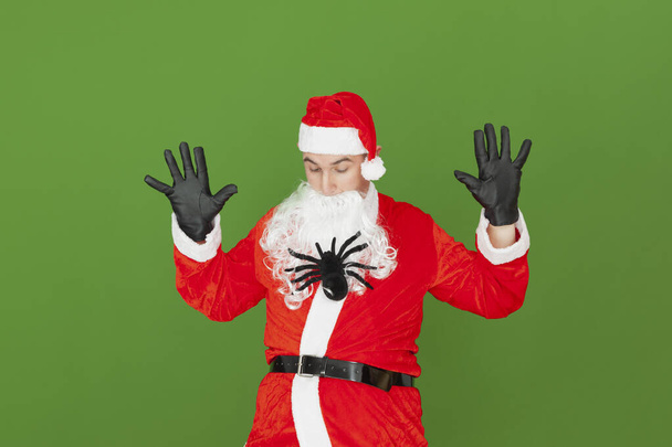 Una araña de juguete está trepando por la barba de un hombre caucásico adulto vestido de Santa Claus, mientras mira asustado. El fondo es verde. - Foto, Imagen