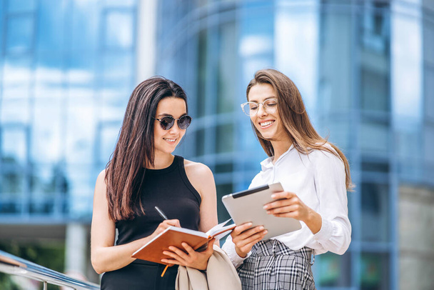 Two young business lady with tablet and notebook walking outdoors near modern business center - Foto, Imagen