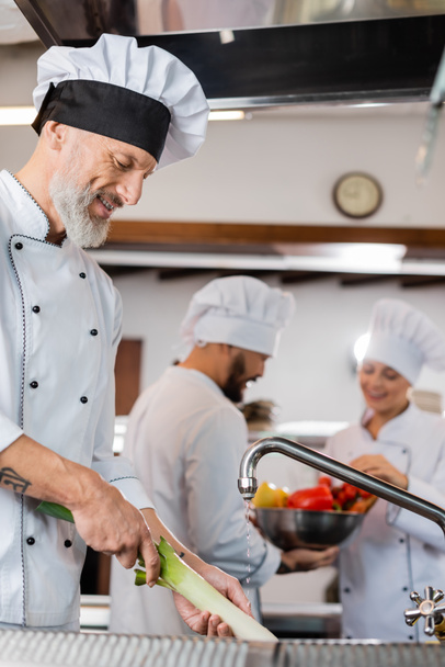 Smiling chef washing leek near blurred interracial colleagues in restaurant kitchen  - Fotó, kép