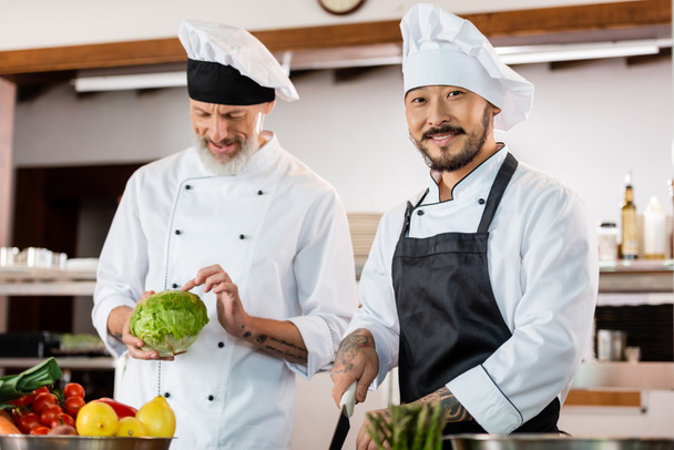 Asian chef with knife smiling at camera near blurred colleague and vegetables in kitchen  - Photo, image