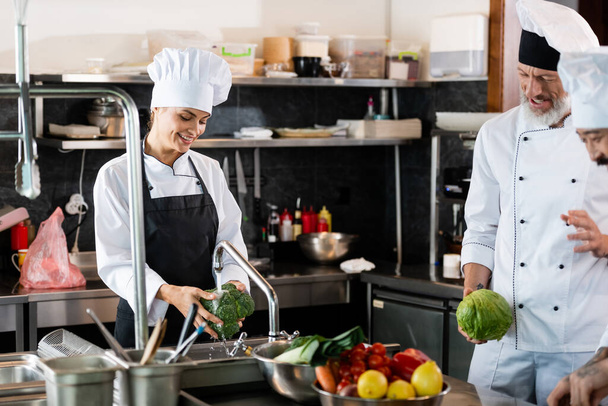Glimlachende chef-kok wassen broccoli in de buurt van interraciale collega 's praten in restaurant keuken  - Foto, afbeelding