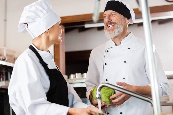 Positive chef holding cabbage near colleague and faucet in kitchen  - Foto, afbeelding