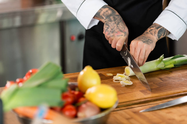 Cropped view of tattooed chef cutting leek near blurred vegetables in kitchen  - Photo, Image