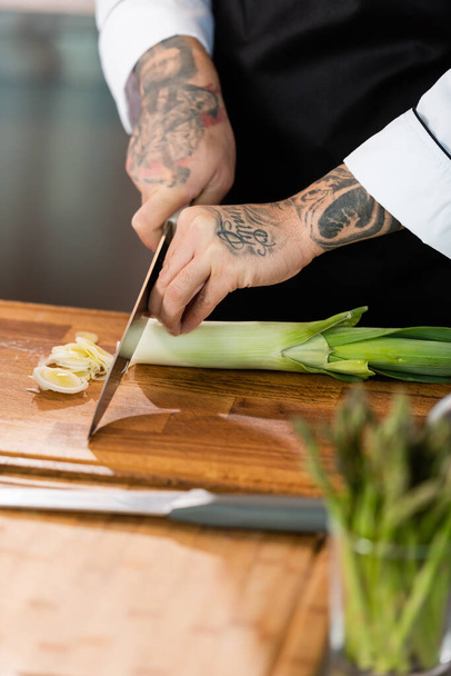 Cropped view of tattooed chef cutting leek near asparagus on blurred foreground in kitchen  - Zdjęcie, obraz