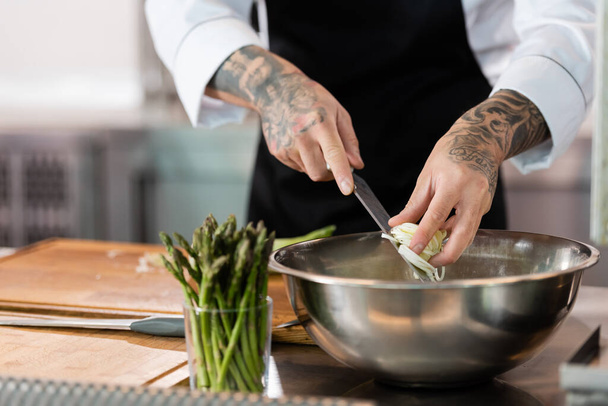 Cropped view of tattooed chef holding knife and sliced leek in kitchen  - Foto, afbeelding