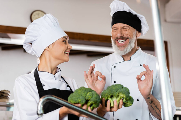 Chef sonriente mostrando buen gesto cerca de colega con brócoli en la cocina  - Foto, Imagen