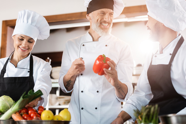 Chef pointing at bell pepper near smiling asian colleague in restaurant kitchen  - Foto, afbeelding