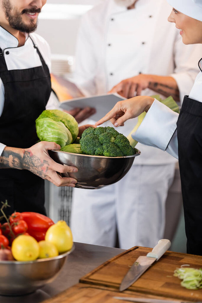 Smiling chef pointing at vegetables in bowl near colleague in kitchen  - Foto, afbeelding