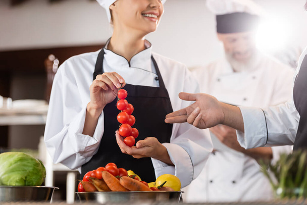 Chef pointing at cherry tomatoes near smiling colleague in restaurant kitchen  - Valokuva, kuva
