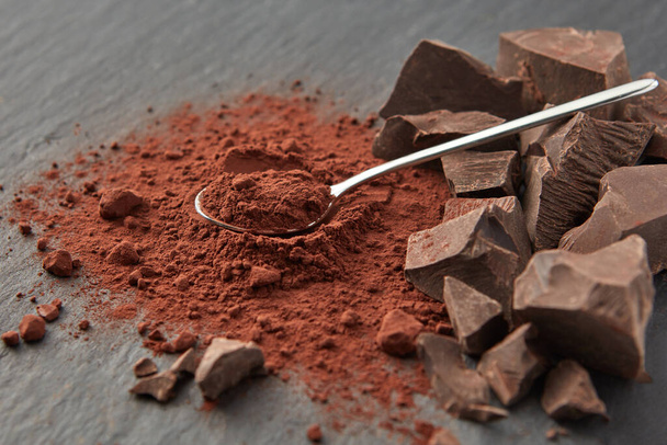 Arranged shiny teaspoon on pile of dry cocoa powder and heap of organic chocolate pieces on gray surface - Φωτογραφία, εικόνα