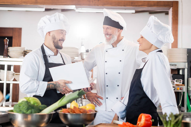 Asian chef holding cookbook and talking to smiling colleagues near food in kitchen  - Foto, imagen