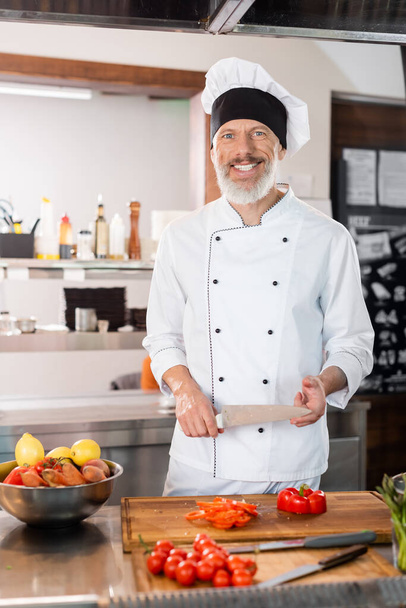 Glimlachende chef met mes in de buurt van rijpe groenten en snijplanken in de keuken  - Foto, afbeelding