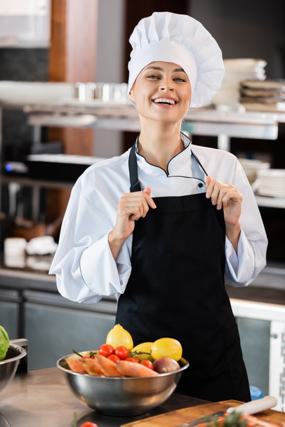 Позитивный шеф-повар трогает фартук и смотрит на камеру рядом со свежими овощами на кухне  - Фото, изображение