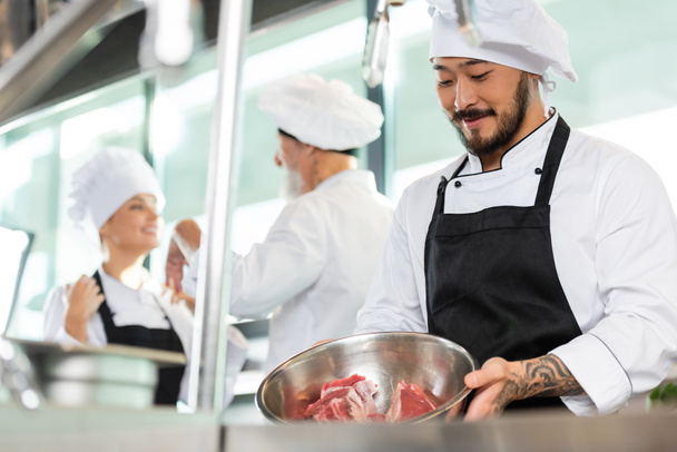 Улыбающийся азиатский шеф-повар держит миску с мясом рядом с коллегами на кухне  - Фото, изображение