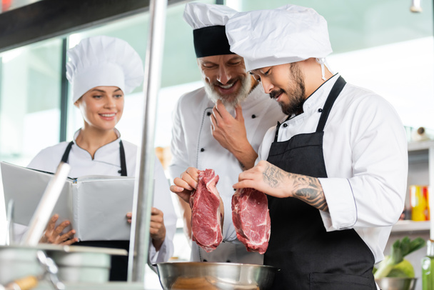 Asian chef holding raw meat near smiling colleagues with cookbook in kitchen  - Foto, Imagen