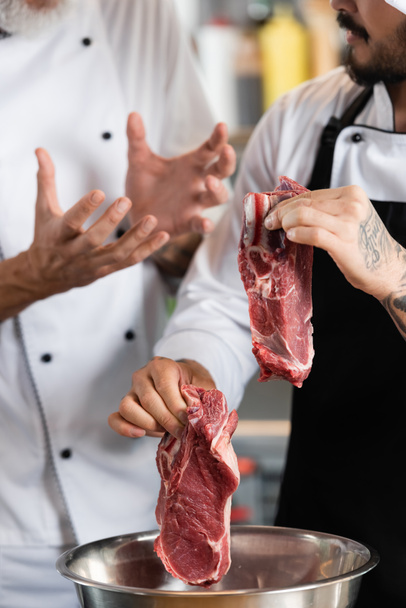 Обрезанный вид на шеф-повара, держащего сырое мясо возле миски и коллегу на кухне  - Фото, изображение