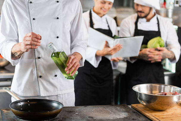 Chef mit Flasche Olivenöl in der Nähe der Pfanne und verschwommene Kollegen mit Kochbuch in Küche  - Foto, Bild