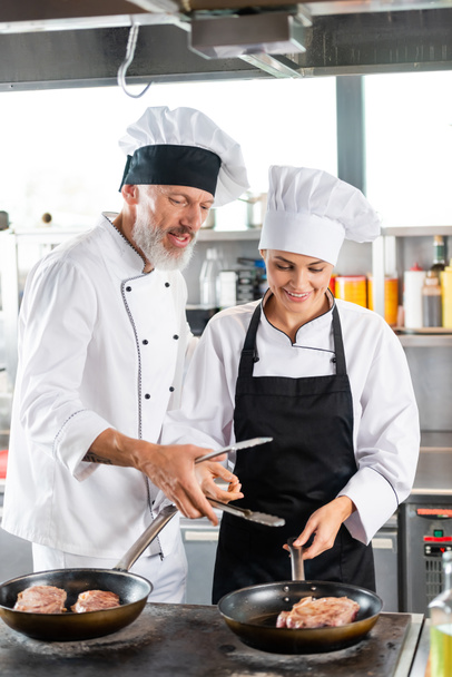 Шеф-повар держит щипцы рядом с улыбающимся коллегой и мясо на сковородках на кухне  - Фото, изображение