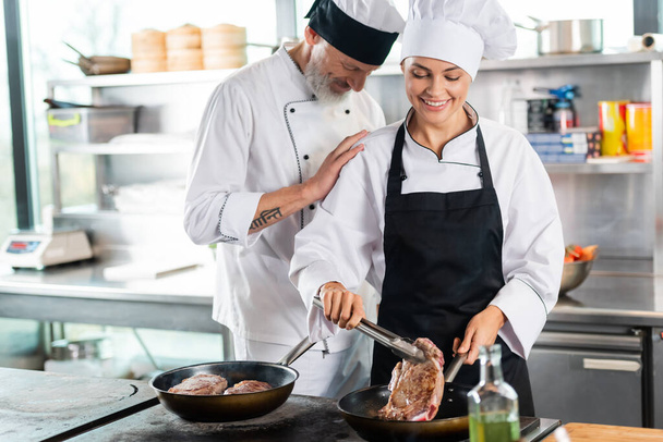 Chef standing near smiling colleague roasting meat in kitchen  - Photo, Image