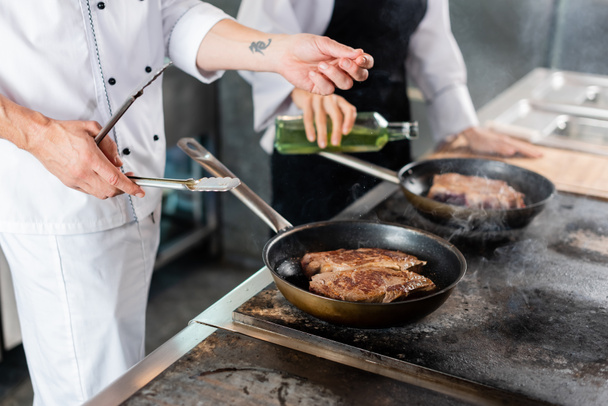 Καλλιεργημένη άποψη του σεφ κρατώντας λαβίδες, ενώ ψήσιμο κρέατος σε βάση εστιών στην κουζίνα  - Φωτογραφία, εικόνα