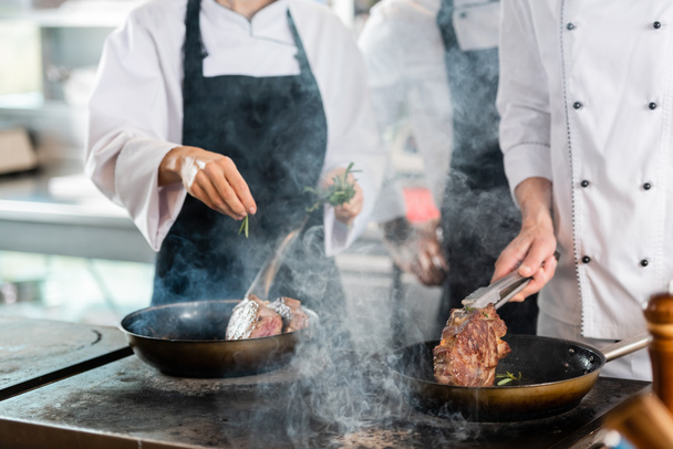 Обрезанный вид шеф-поваров с щипцами и розмарином жарки мяса на кухне  - Фото, изображение
