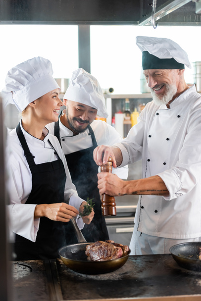 Усміхнений шеф-кухар приправляє м'ясо на сковороді біля веселих багатоетнічних колег з розмарином на кухні
  - Фото, зображення