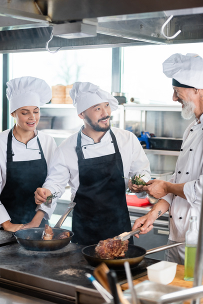 Asian chef holding rosemary near smiling colleagues cooking meat in kitchen  - Φωτογραφία, εικόνα