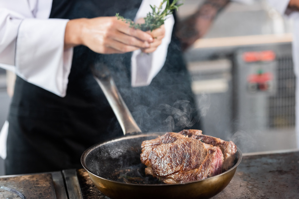 Roasted meat on frying pan near blurred chef holding rosemary in kitchen  - Photo, image