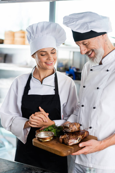 Smiling chef holding roasted meat on cutting board near colleague in apron  - Фото, зображення