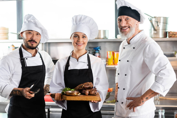 Smiling chef holding roasted meat on cutting board near interracial colleagues in kitchen  - Foto, imagen