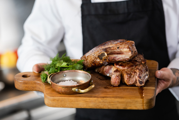 Cropped view of blurred chef holding roasted meat and greens on cutting board in kitchen  - Φωτογραφία, εικόνα