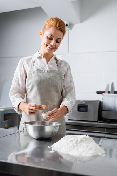 Chef sonriente sosteniendo huevo cerca de harina y tazón en la cocina  - Foto, Imagen