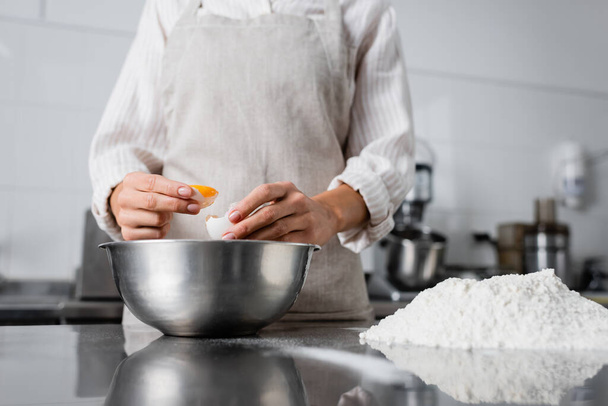 Oříznutý pohled na šéfkuchaře držícího vejce v blízkosti misky a mouky na kuchyňském stole  - Fotografie, Obrázek