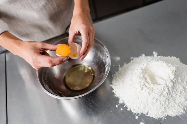 Cropped view of chef holding egg and eggshell near bowl and flour on table  - Foto, Bild