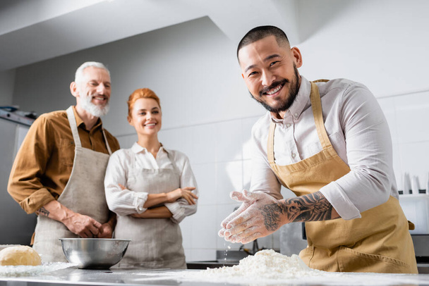 Positive asian chef in apron standing near flour and blurred colleagues in kitchen  - Foto, Bild