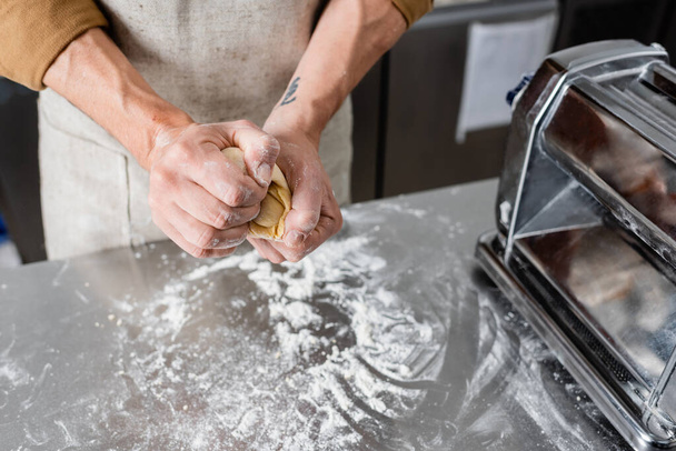 Vista recortada del chef sosteniendo masa cerca de la máquina de hacer pasta y harina en la mesa  - Foto, Imagen