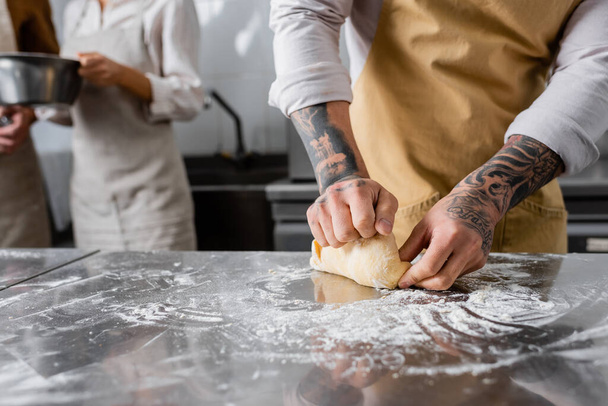 Oříznutý pohled na tetovaného kuchaře, jak dělá těsto poblíž rozmazaných kolegů v kuchyni  - Fotografie, Obrázek
