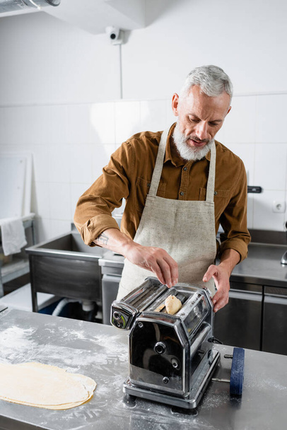 Зрелый шеф-повар кладет тесто в машину для приготовления макарон на кухне  - Фото, изображение