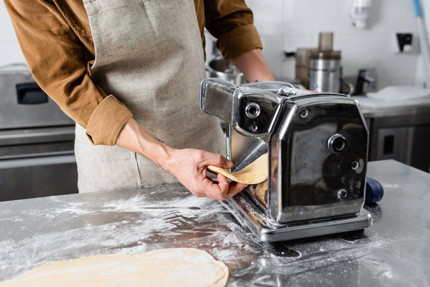 Vista recortada del chef en delantal sosteniendo la masa cerca de la máquina de hacer pasta y harina en la cocina  - Foto, imagen