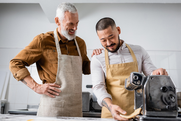 Smiling chef in apron standing near asian colleague holding dough near pasta maker machine in kitchen  - Valokuva, kuva