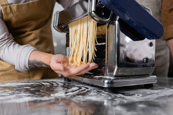 テーブルの上に小麦粉の近くのパスタメーカーのマシン上でスパゲティを作る入れ墨シェフの作物ビュー  - 写真・画像