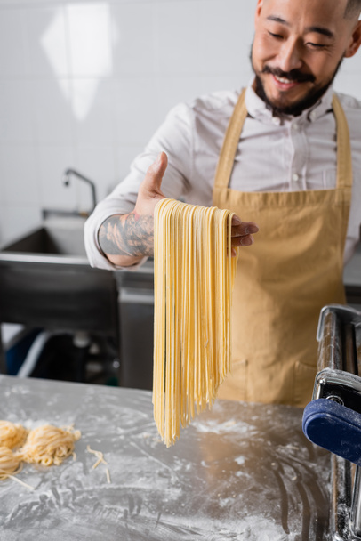 Blurred asian chef holding raw spaghetti near flour and pasta maker machine in kitchen  - Fotoğraf, Görsel