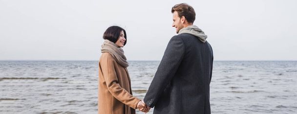 pareja complacida en abrigos de otoño tomados de la mano y mirándose mientras caminan cerca del mar, pancarta - Foto, Imagen