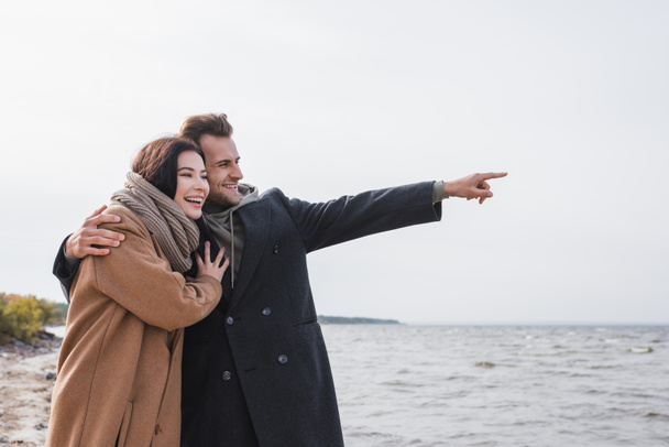happy man pointing with finger while hugging girlfriend near sea - Foto, afbeelding