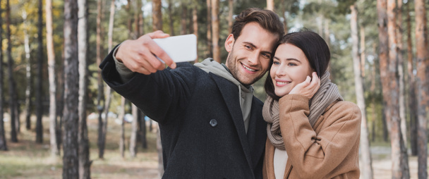 smiling man and woman in autumn coats taking selfie on smartphone in park, banner - Φωτογραφία, εικόνα